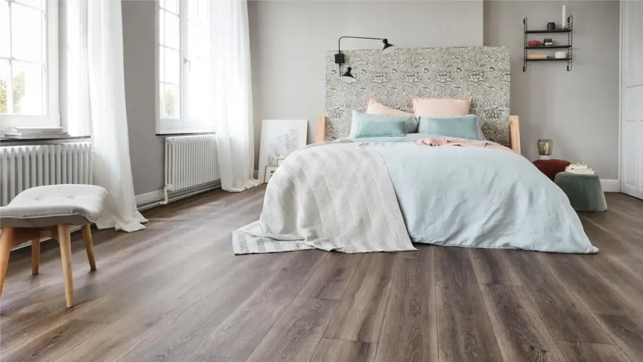 Un lit douillet avec un couvre-lit ray&eacute; et des coussins bleus et roses, sur un rev&ecirc;tement sol chambre imitation bois sombre.