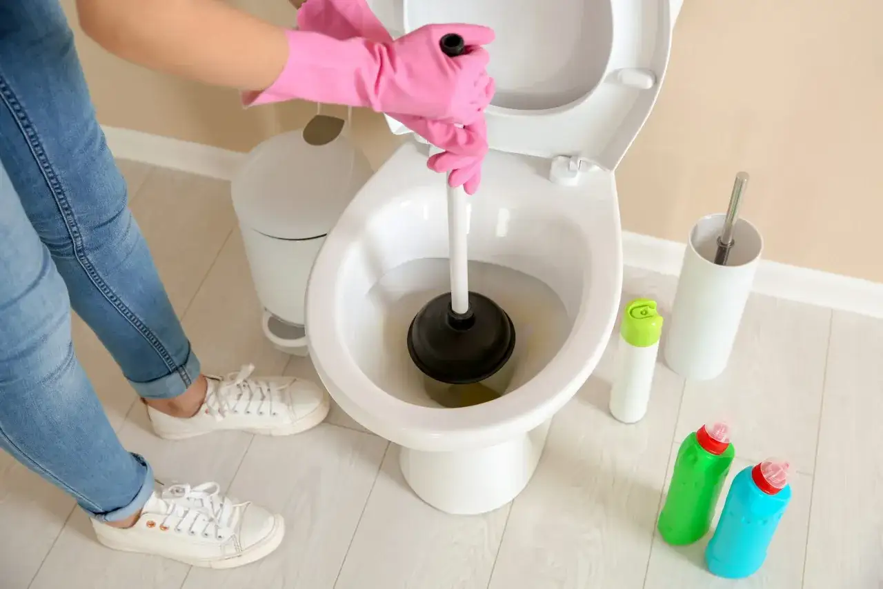 Personne avec des gants roses utilise une ventouse pour déboucher un WC. Des produits de nettoyage sont à proximité.