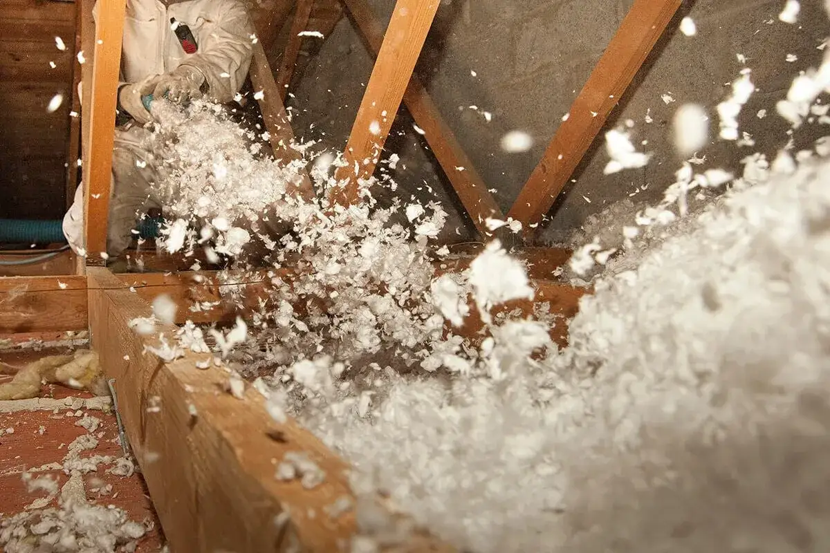 Un ouvrier en combinaison souffle de l'isolant dans des combles. L'isolation des combles par soufflage est en cours, créant un nuage de flocons blancs.
