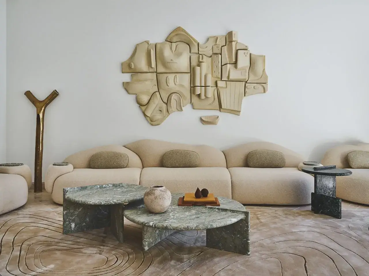 Une sculpture abstraite en terre cuite vient habiller un mur blanc, dominant un canapé beige aux formes douces et des tables basses en marbre vert.