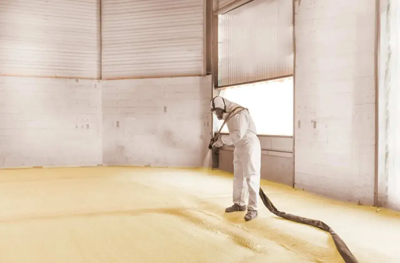 Un ouvrier en combinaison intégrale applique du polyuréthane projeté sur le sol d'un entrepôt.