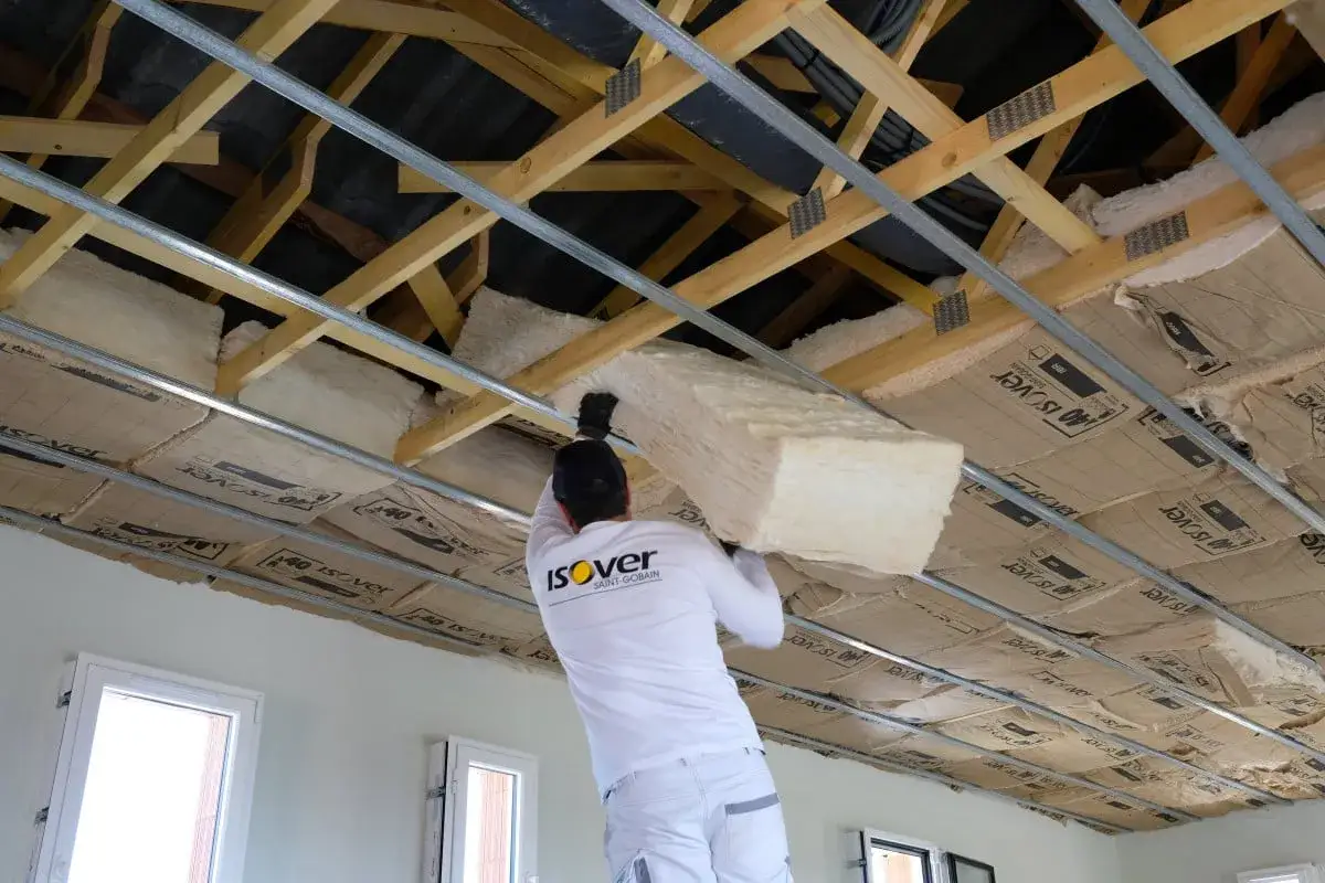 Un ouvrier installe un panneau de fibre de verre pour isoler un plafond. La structure en bois et les rails métalliques sont visibles.