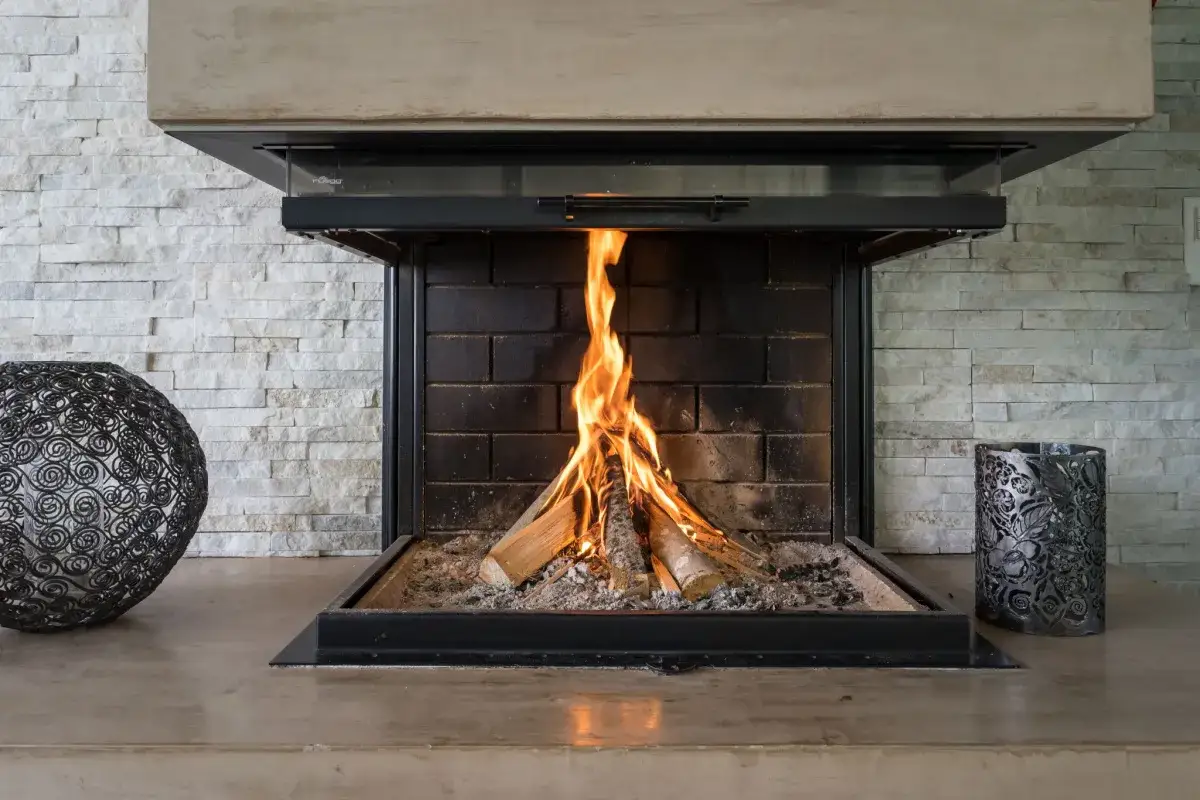 Un feu cr&eacute;pite dans une chemin&eacute;e ouverte, r&eacute;chauffant la pi&egrave;ce. Des d&eacute;corations m&eacute;talliques encadrent le foyer.