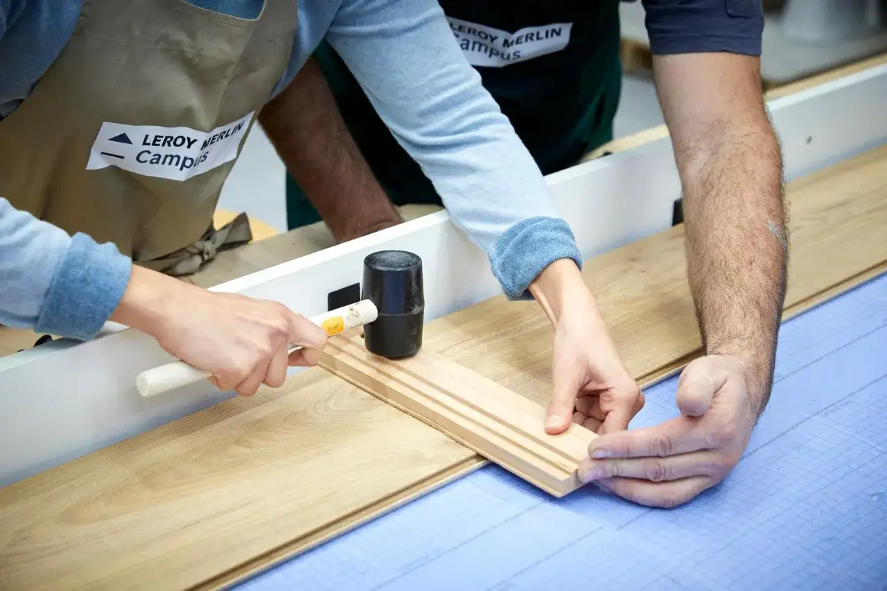 Deux mains assemblent des lames de parquet stratifié. L'une utilise un maillet pour ajuster une pièce, l'autre guide.