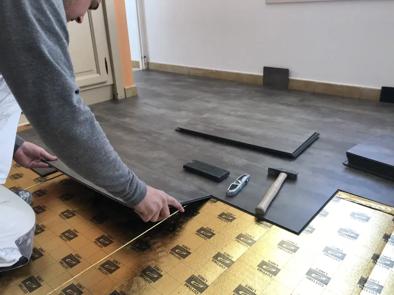 Pose d'une lame PVC clipsable sur une sous-couche dorée. Un marteau et un cutter sont à proximité.
