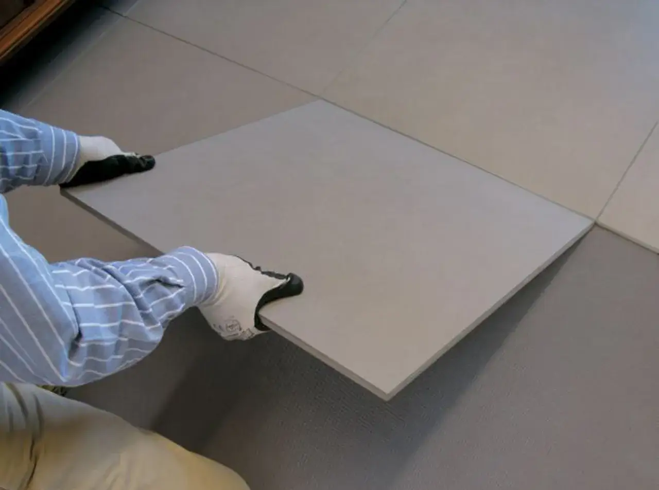 Pose d'une nouvelle dalle pour carreler sur du carrelage existant. Mains gantées tenant une grande dalle grise.