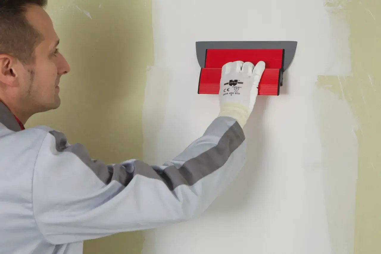 Un ouvrier en combinaison grise utilise un outil rouge pour lisser du pl&acirc;tre sur un mur. La question 