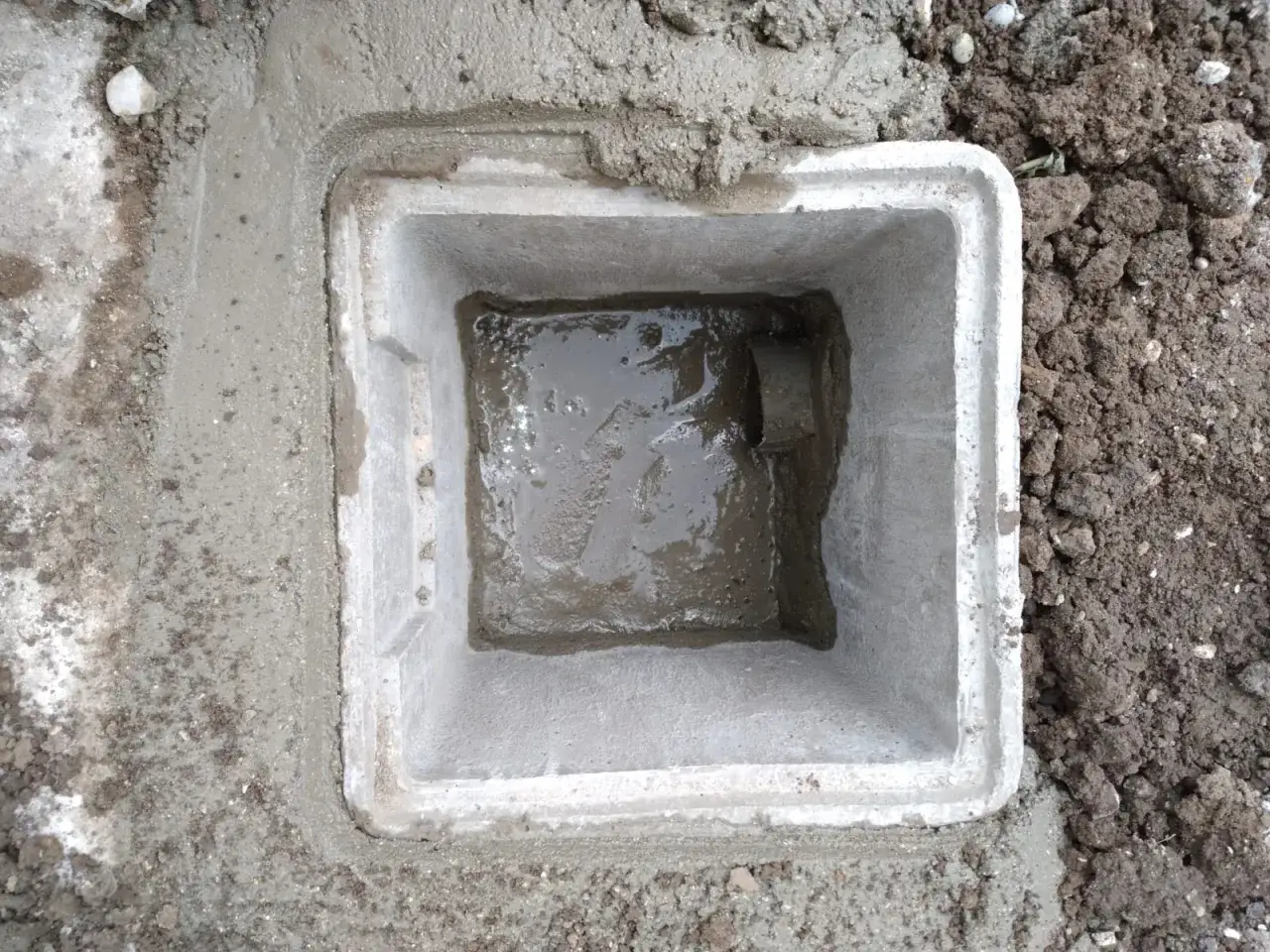 Un regard de béton fraîchement posé, rempli d'eau pluviale. Le sol environnant est meuble et terreux.