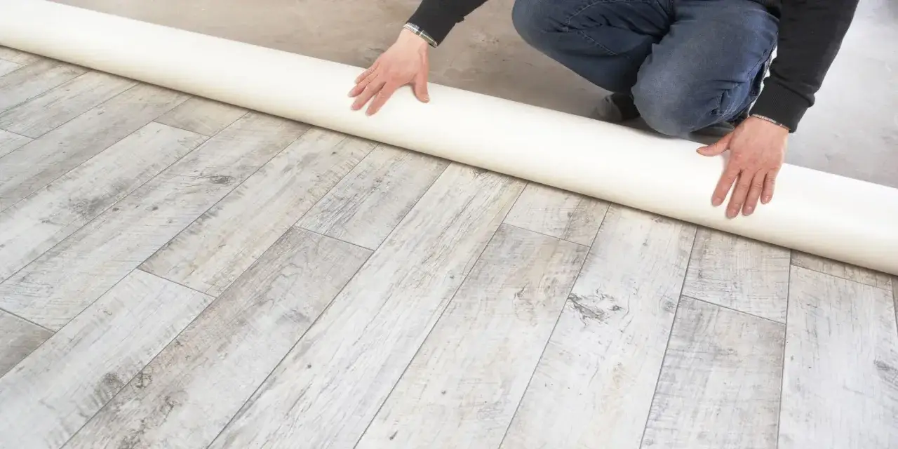 Un rouleau de pose lino est d&eacute;roul&eacute; sur un sol imitation bois vieilli. Les mains guident le mat&eacute;riau.