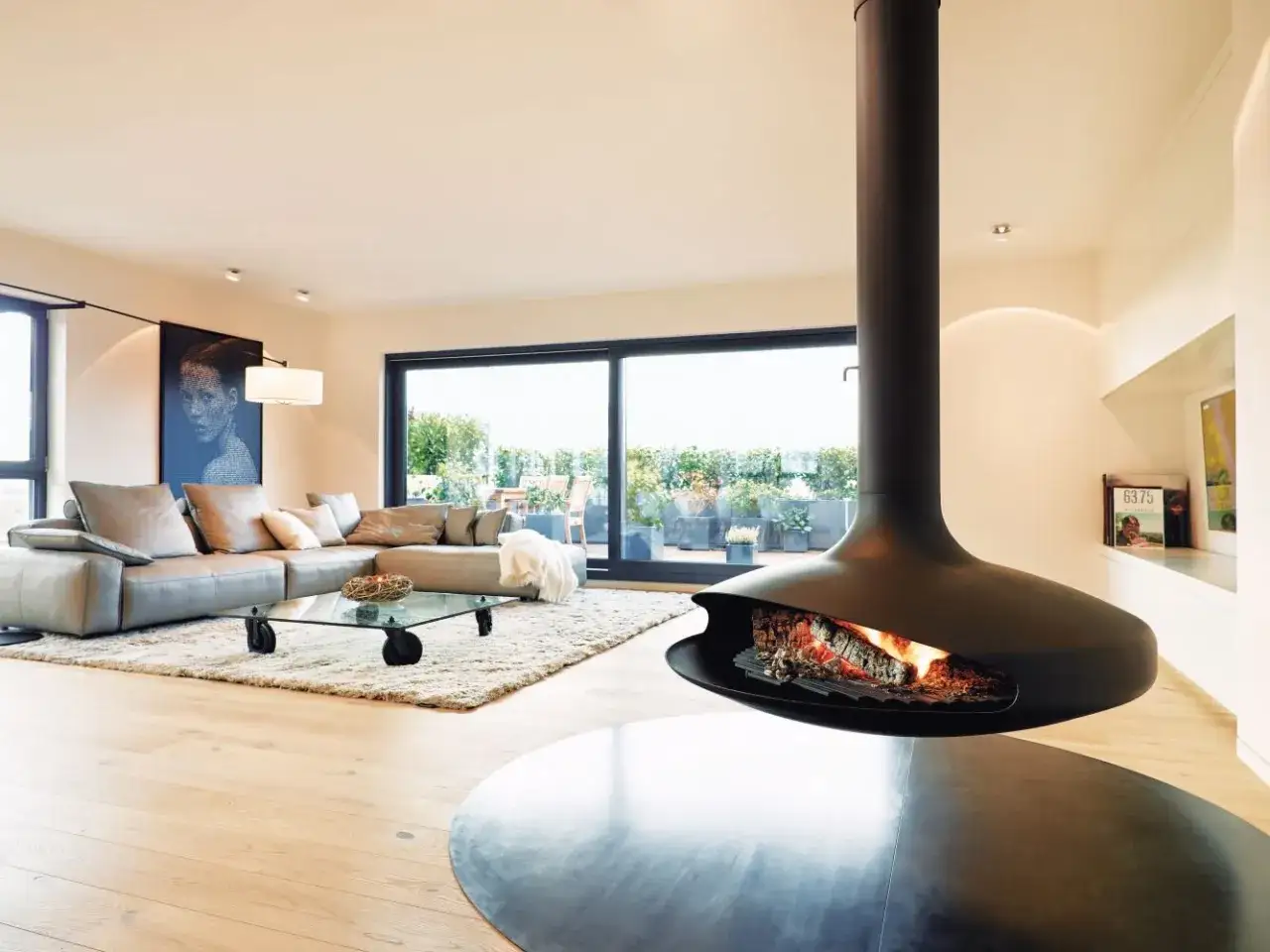 Un salon moderne avec une chemin&eacute;e suspendue noire, un canap&eacute; gris, une table basse en verre et une grande baie vitr&eacute;e donnant sur une terrasse. Id&eacute;al pour moderniser une chemin&eacute;e.