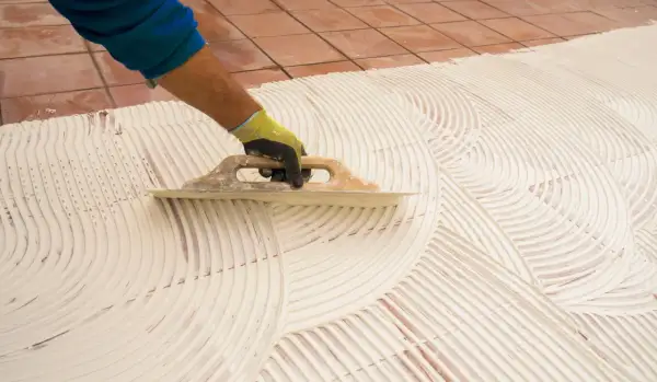 Main tenant, pose carrelage 60x60. Un artisan étale la colle avec une truelle pour préparer la pose de grands carreaux.