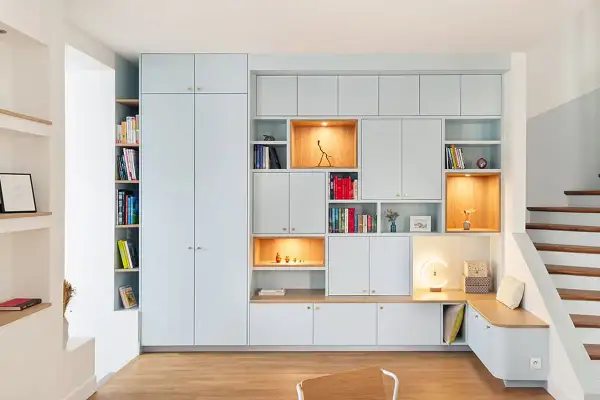 Un mur habillé de rangements modulables bleu pâle, avec des niches éclairées et une banquette intégrée près d'un escalier en bois.
