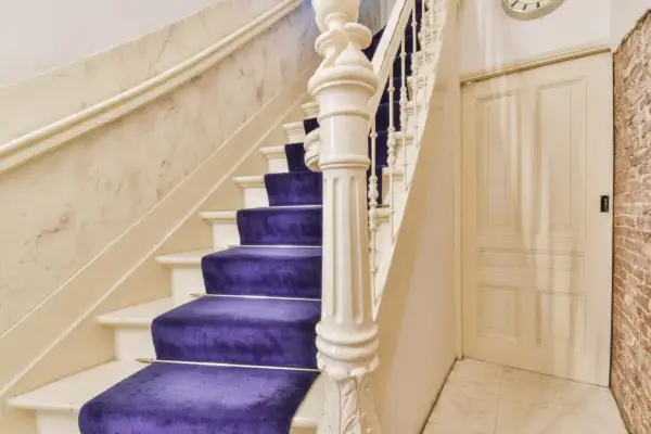 Escalier majestueux avec une moquette violette éclatante, un garde-corps blanc sculpté et un mur en marbre. Une porte crème et un mur de briques complètent la scène.