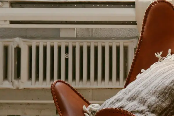 Un fauteuil en cuir marron avec un plaid douillet est placé près de vieux panneaux rayonnants blancs, créant une ambiance chaleureuse.