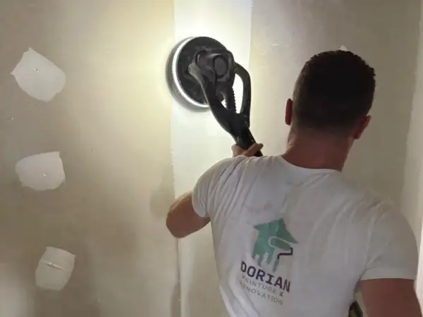 Un ouvrier utilise une ponceuse pour lisser un mur, préparant la surface pour poser bande placo.