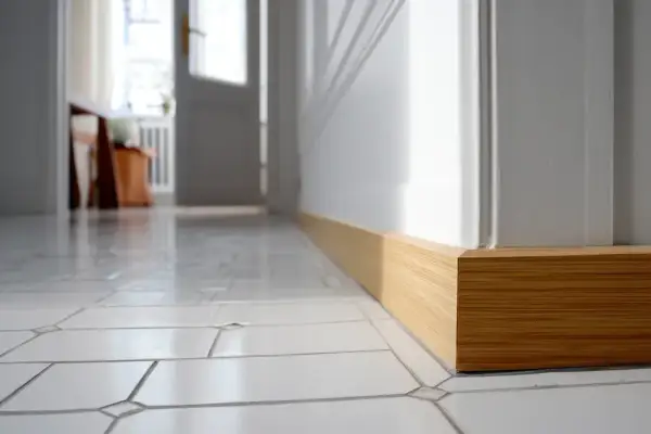 Pose de plinthe en bois clair, finition impeccable sur un sol carrelé blanc. La lumière du jour met en valeur le travail.
