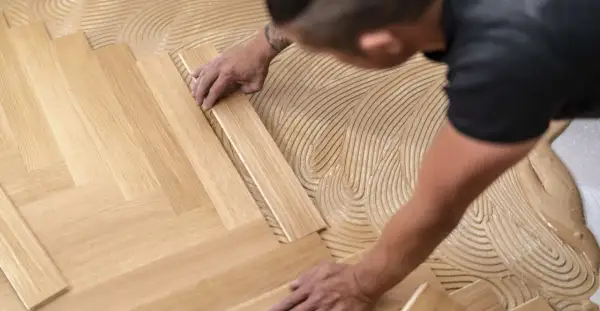 Pose d'un parquet flottant en chevrons. Un artisan applique la colle sur le sol avant de placer les lames de bois.