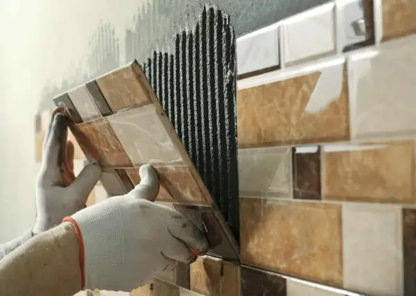 Mains gantées appliquant du mortier pour la pose carrelage d'une mosaïque murale aux tons marron et beige.