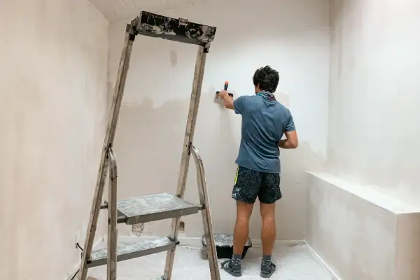 Un homme enlever du crépi intérieur sur un mur, à l'aide d'une spatule, près d'un escabeau.