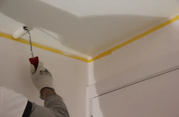Main tenant en main un rouleau, il s'apprête à peindre un plafond. Le ruban de masquage jaune protège les bords.