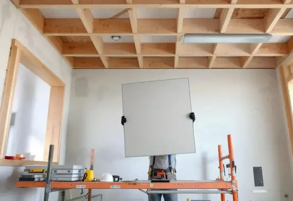 Un ouvrier sur un échafaudage s'apprête à poser une plaque pour faire un faux plafond. La structure en bois est visible.