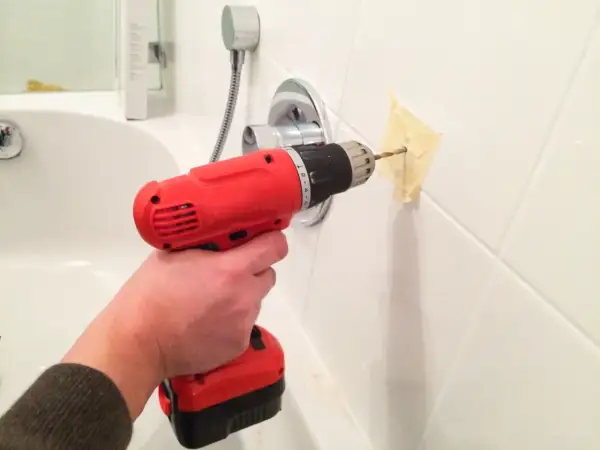 Une main utilise une perceuse pour percer du carrelage dans une salle de bain, avec du ruban adhésif pour guider le foret.