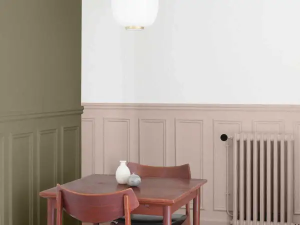 Peinture salon deux tons : mur vert olive contrastant avec un soubassement rose poudré orné de moulures. Une table en bois et des chaises sont disposées près d'un radiateur.